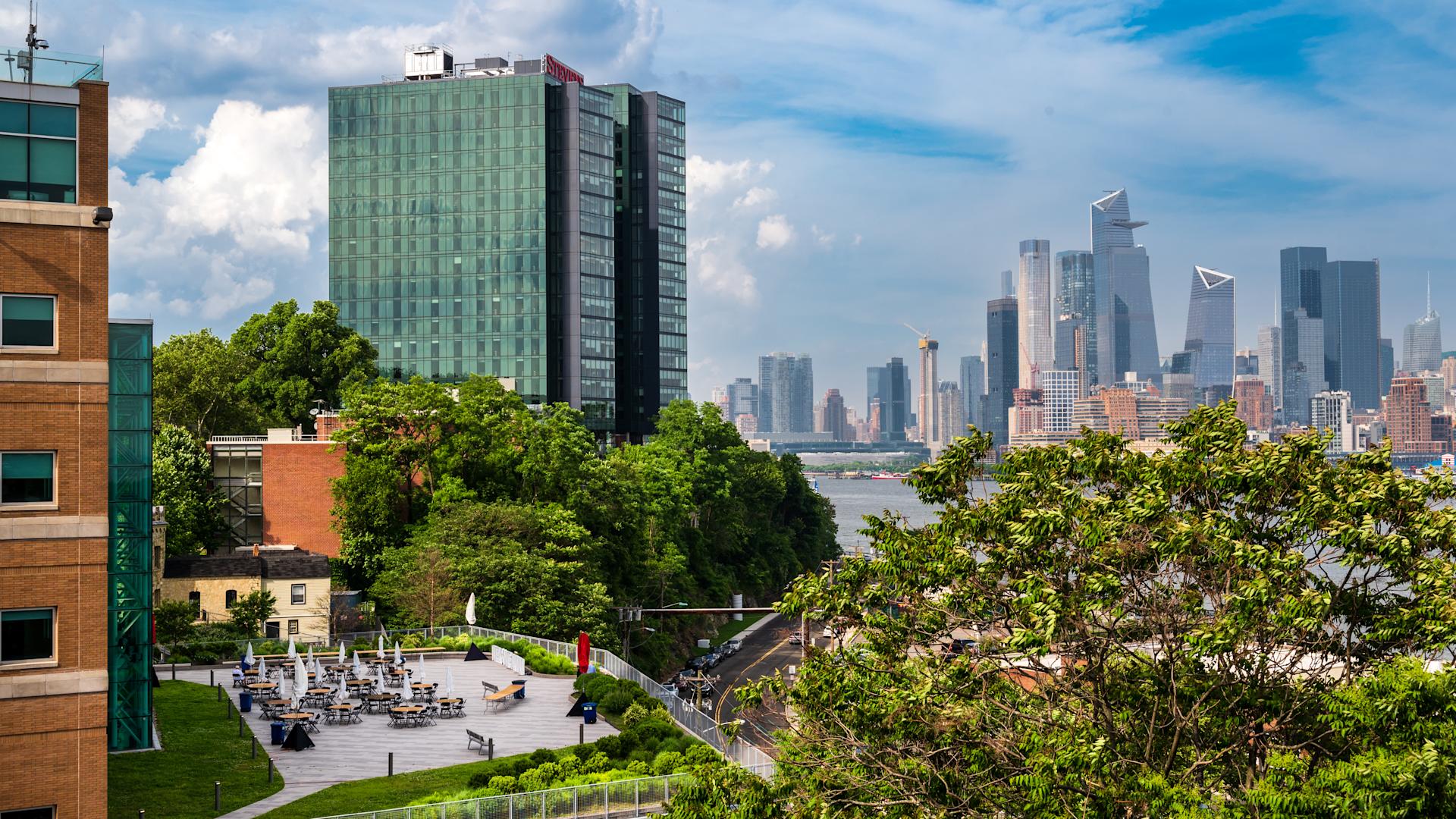 Campus terrace with café tables and umbrellas in summer, the Midtown Manhattan skyline beyond the river