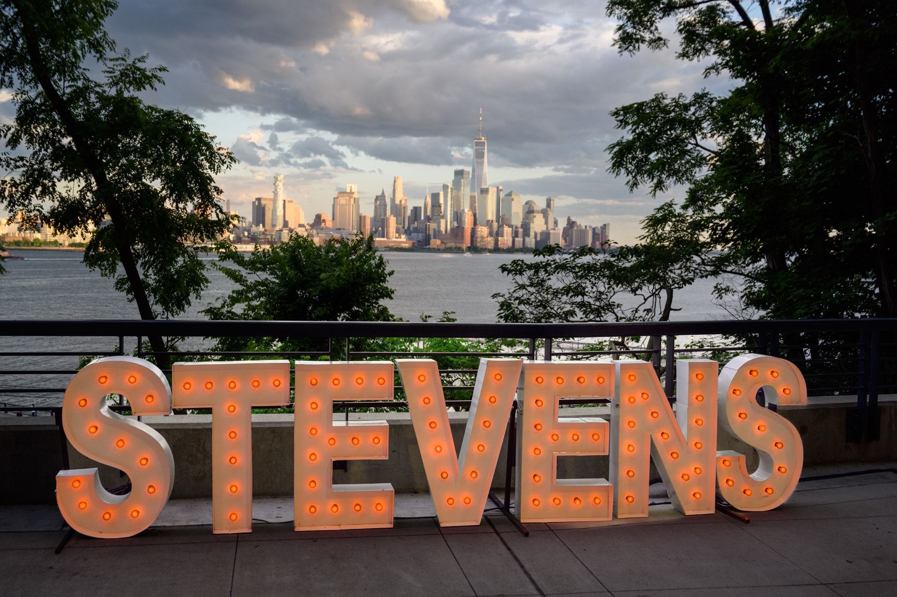 Illuminated STEVENS marquee letters on the campus overlook at sunset, with the lower Manhattan skyline behind
