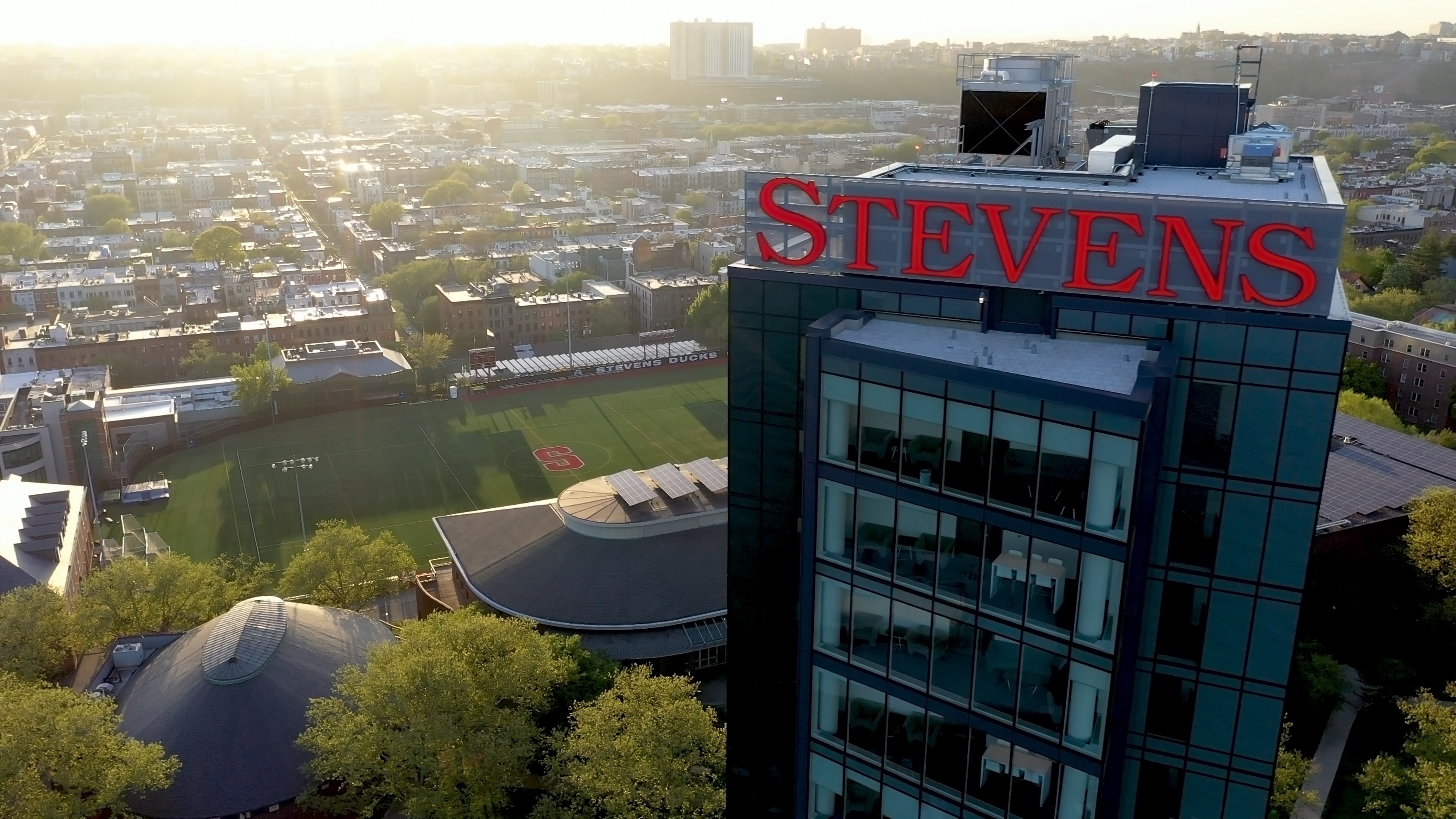 Sunrise aerial of the campus rooftop sign above the athletic field and the Hoboken street grid
